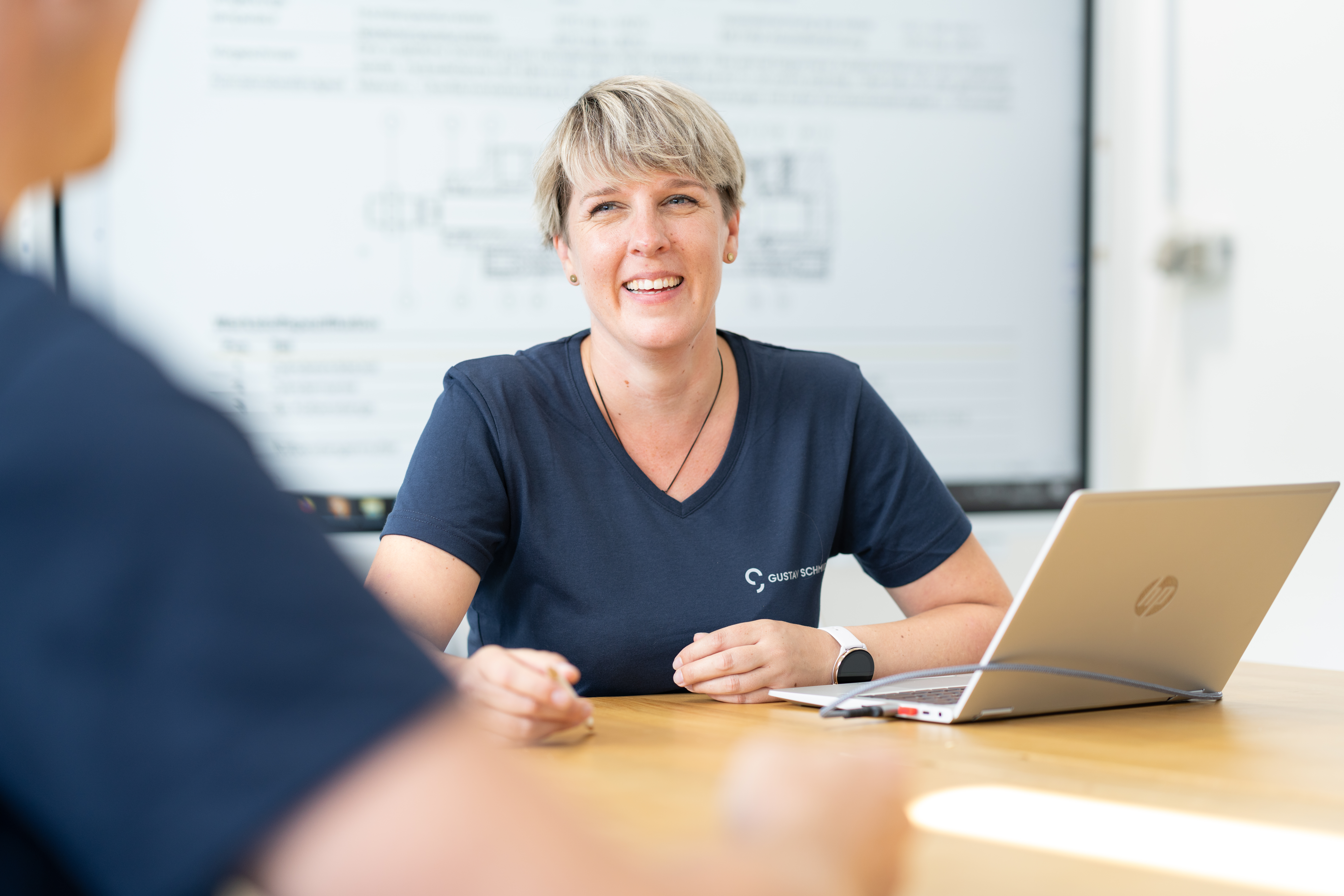 Frau mit blonden Haaren und blauem Gustav-Schmidt-Shirt schaut andere Person (nur verschwommen von hinten sichtbar, vor ihr sitzend) mit lachendem Gesicht an, im Hintergrund digitale Präsentationstafel zu sehen, am Tisch vor ihr ein Laptop und hat Stift in der Hand